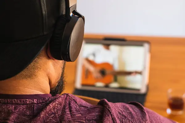 Şapka ve kulaklık takan iri yarı, sakallı bir adam tableti aracılığıyla evde gitar dersi alıyor. Seçici odak.