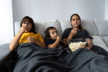 Beyaz anne ve kızları oturma odasındaki kanepede oturup patlamış mısır yerken film izliyorlar.,