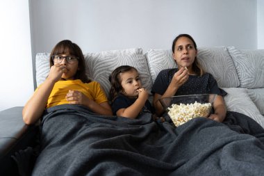 Beyaz anne ve kızları oturma odasındaki kanepede oturup patlamış mısır yerken film izliyorlar.,