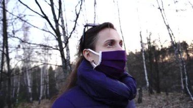 Korunaklı maskeli bir kız güneşli bir ormanda Coronavirus 'a karşı genç sarı bir çiçeğin baharını koklamak ister.