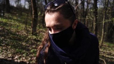 Korunaklı maskeli bir kız güneşli bir ormanda Coronavirus 'a karşı genç sarı bir çiçeğin baharını koklamak ister.