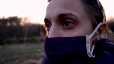Korunaklı maskeli bir kız güneşli bir ormanda Coronavirus 'a karşı genç sarı bir çiçeğin baharını koklamak ister.