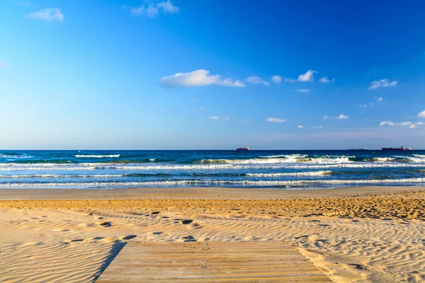 Deck de madeira sobre praia de areia com céu azul e oceano no fundo. Espuma branca no topo das ondas do oceano em Tarragona Espanha — Fotografia de Stock