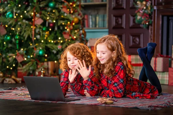 Funny red-haired children next to the Christmas tree and gifts communicate online through a laptop.