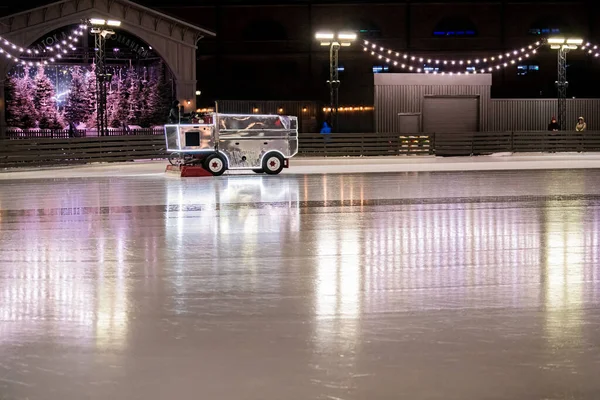 On an open ice skating rink, lit with festive lights and garlands, a combine recovers ice for ice skating.