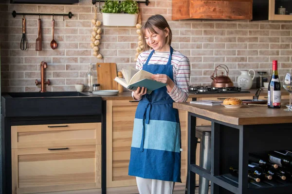Happy woman in a linen apron standing in the kitchen at the table with a smile leafing through a cookbook