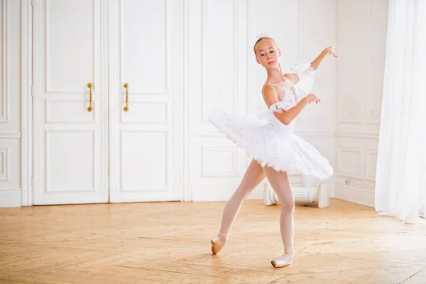 young slender ballerina in a white tutu standing on pointe at a ballet bar in a beautiful white hall in front of a mirror.