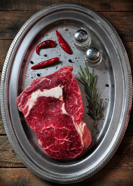 Bife de vaca suculento cru com alecrim, sal, pimenta e pimenta em um prato de ferro na mesa de madeira — Fotografia de Stock