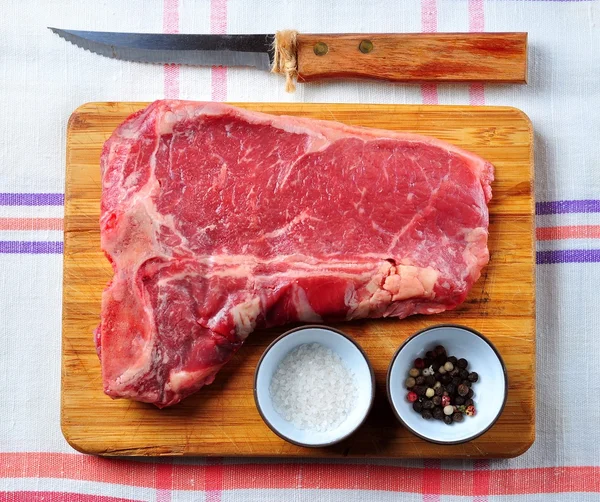 Bife suculento cru de carne de T-Bone em uma placa de madeira com sal, pimenta e uma faca — Fotografia de Stock