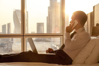Young businessman using smartphone and laptop in bed