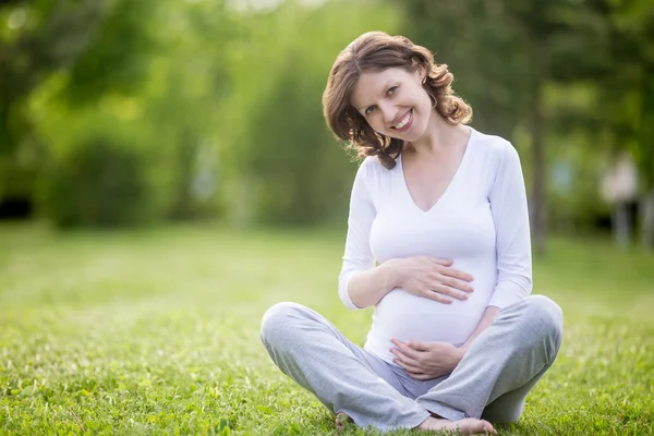 Sonriendo futura mamá sentado con las piernas cruzadas en el césped de hierba —  Fotos de Stock