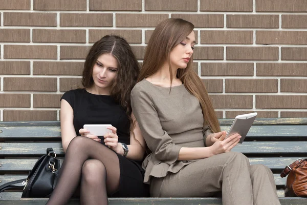 Dos mujeres jóvenes sentadas en el banco usando sus propios dispositivos — Foto de Stock