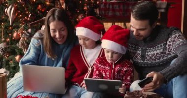Elektronik aletler kullanan çocuklu aileler Noel ağacının yanına oturur.
