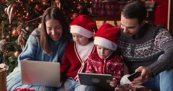 Familia con niños usando dispositivos electrónicos se sientan cerca del árbol de Navidad — Vídeo de stock