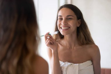 Güzel, genç bir bayan, parlak bir gülümsemeyle banyo aynasında duruyor.