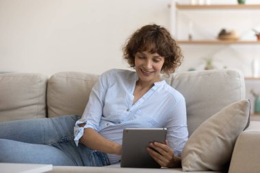 Kapatın, gülümseyen genç bayan kanepede yatıyor, tablet kullanıyor.