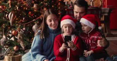 Aile, süslü Noel Ağacı 'nın yanında oturup akıllı telefon kullanarak eğleniyor.