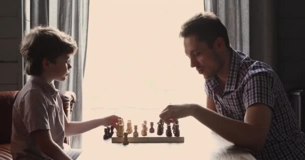 Little son and his young father play chess seated indoor — Stock Video