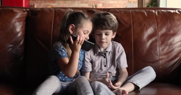 Τα παιδιά κάθονται στον καναπέ στο σπίτι διασκεδάζοντας χρησιμοποιώντας το smartphone — Αρχείο Βίντεο