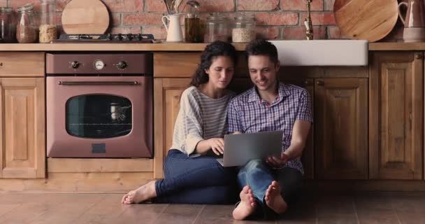 Pareja casada descansando sentada en el suelo en cocina con portátil — Vídeo de stock
