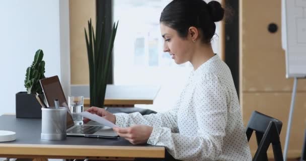 Konzentrierte indische junge Frau hält Papierdokumente in der Hand und arbeitet am Computer. — Stockvideo