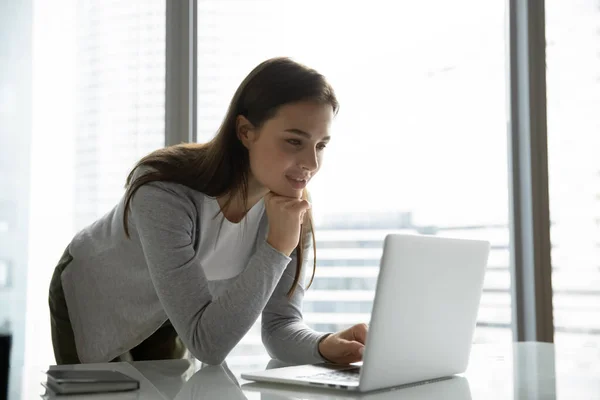 Interessert kvinnelig arbeider som står i nærheten av skrivebordet og ser på PC-skjermen – stockfoto
