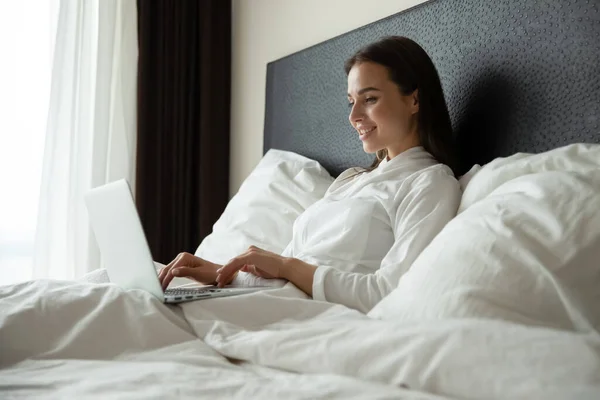 Happy young female remote employee using laptop in comfortable bed