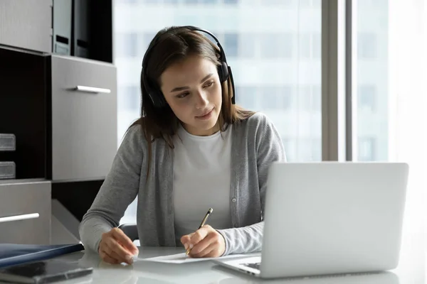 Zainteresovaná žena zaměstnanec sledování webinář ve sluchátkách psaní informací — Stock fotografie