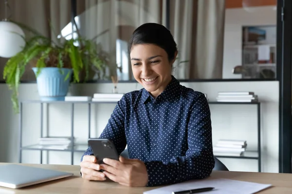 Smiling Indian businesswoman looking at phone screen, sitting in office