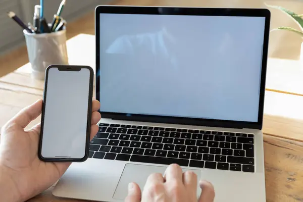 Hombre usando la computadora que sostiene el teléfono inteligente con la pantalla de maqueta vacía blanca — Foto de Stock