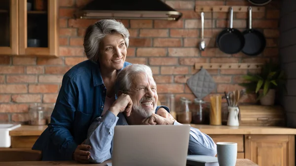 Couple mature rêveuse étreignant, regardant de côté, en utilisant un ordinateur portable ensemble — Photo