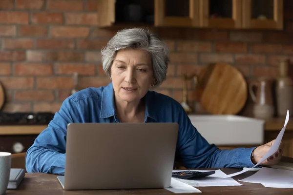 Улыбающаяся взрослая женщина с ноутбука, проверяющая финансовые документы дома — стоковое фото