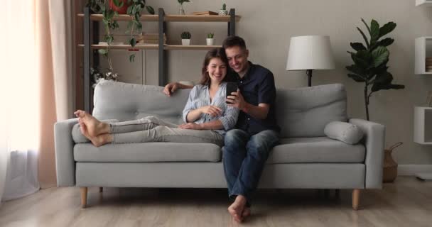 Pasangan Milenial santai di sofa menggunakan smartphone Stok Video Bebas Royalti