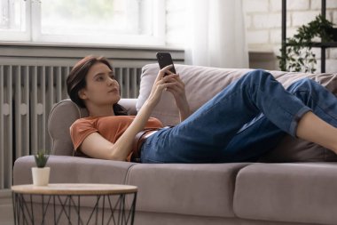 Rahat, sakin, genç bir kadın kanepeye uzanmış, mobil uygulamalar kullanıyor..