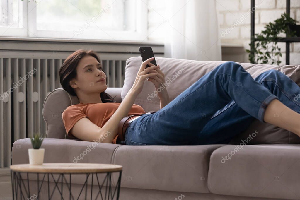 Relaxed calm young woman lying on sofa, using mobile apps.