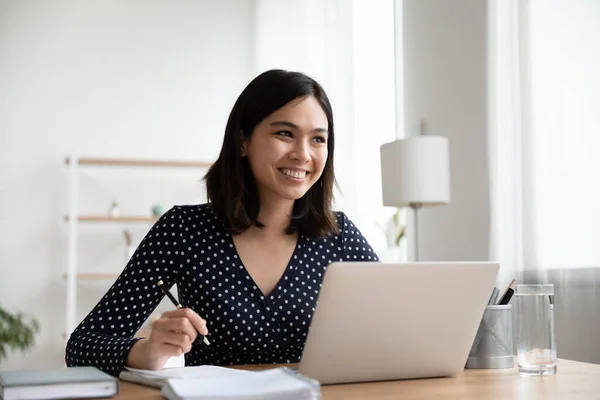 Dreamy smiling Asian businesswoman sitting at desk with laptop — Stock Photo, Image