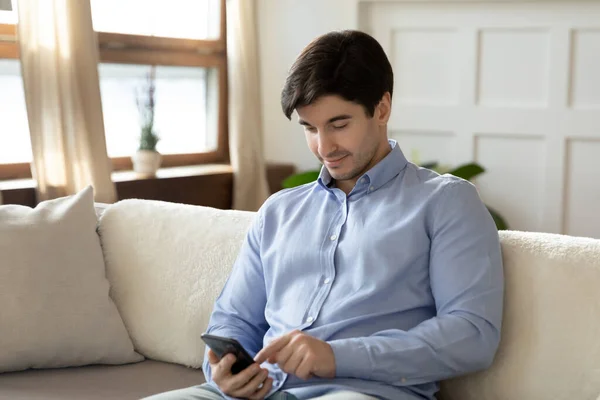 Happy millennial man resting on sofa using smartphone chatting online