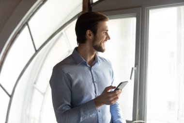 Ofis penceresinin yanında elinde akıllı telefon tutan iyimser genç adam.
