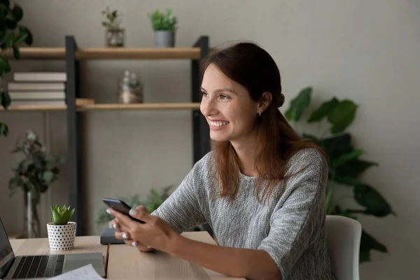 Dreamy young woman looking in distance holding smartphone in hands.