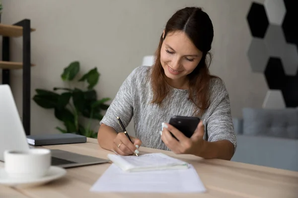 Uśmiechnięta młoda biała kobieta używa smartfona ucząc się w domu. — Zdjęcie stockowe