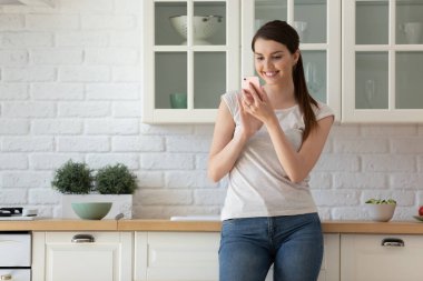 Gülümseyen genç kadın telefonu kullanıyor, modern mutfakta duruyor.