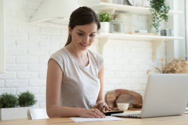 Gülümseyen genç kadın mali durumları kontrol ediyor, laptop kullanıyor, mutfakta oturuyor.