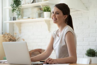 Gülümseyen genç bir kadın dizüstü bilgisayar kullanıyor, faturaları hesaplıyor, mutfakta oturuyor.