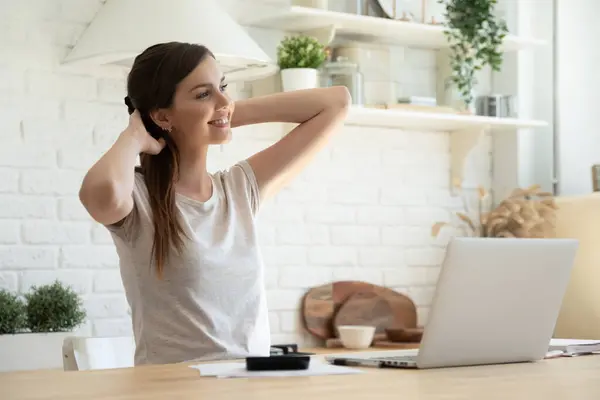 Mujer sonriente estirándose, usando laptop, sentada a la mesa en la cocina —  Fotos de Stock