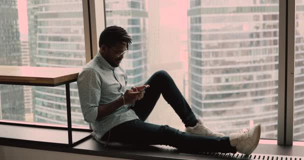 Relaxed african guy seated on windowsill indoor with smartphone — Vídeos de Stock
