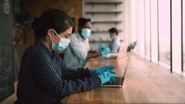 Multiethnic staff wearing facemasks gloves working on laptops keeping distance