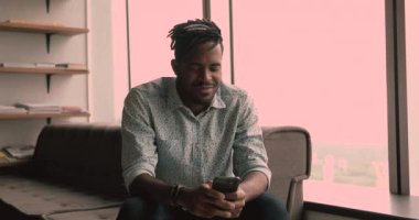 Afrikalı adam akıllı telefon kullanarak modern ofiste oturuyor.