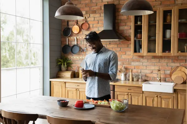 Young happy african american man using smartphone, distracted from cooking. — Stockfoto