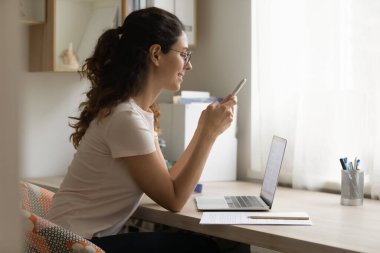 Gözlüklü, gülümseyen bir kadın telefon kullanıyor, çalışma masasında oturuyor.