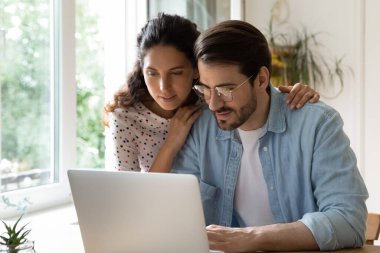 Genç kadın kocasına arkadan sarılıyor, birlikte dizüstü bilgisayar kullanıyor.